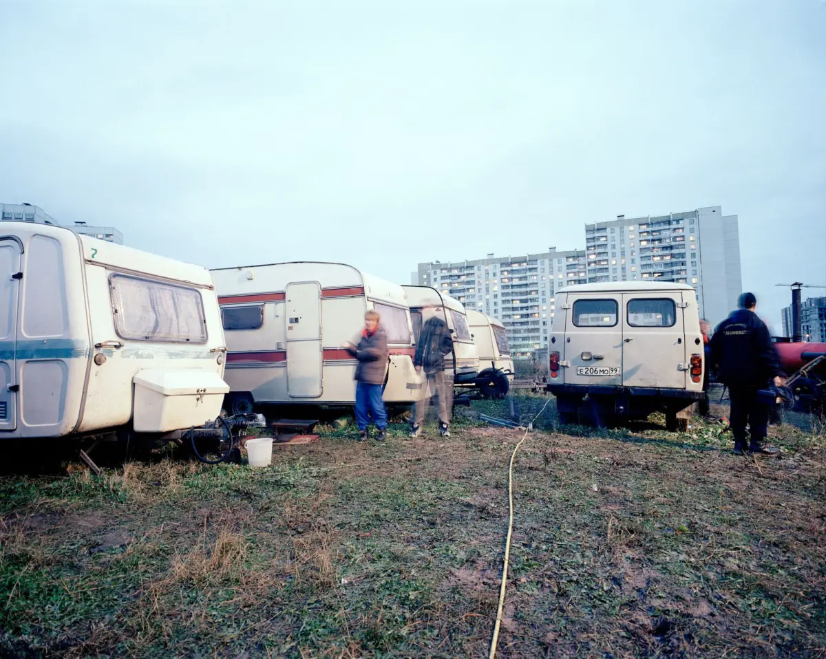 Row of white caravans in a grassy lot, blurred pedestrians nearby, with high-rise apartment buildings in the background.