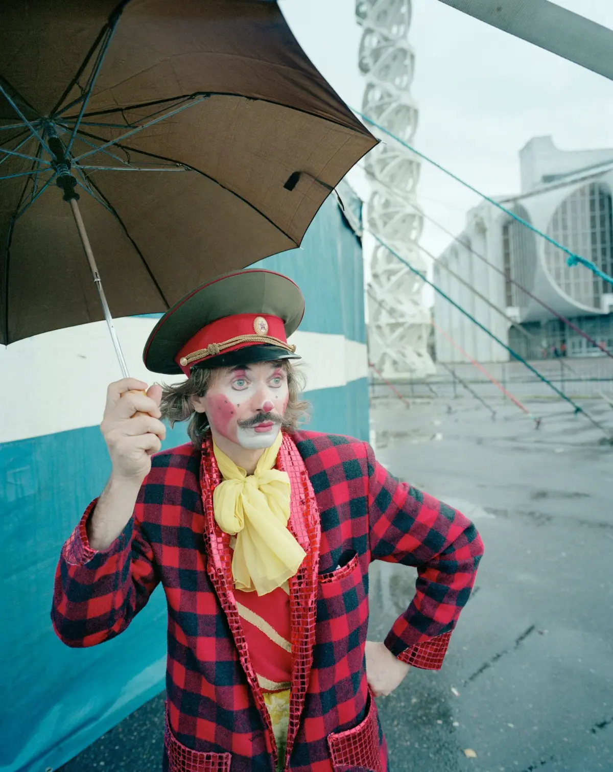 Clown in red plaid suit with yellow scarf and peaked red cap, holding a brown umbrella in a wet urban construction area.