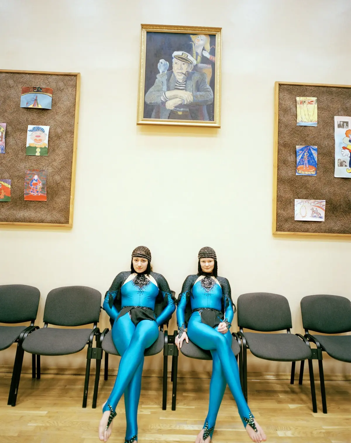 Two people in matching blue metallic bodysuits sit on chairs in a gallery, with framed painting and artwork on corkboards behind them.