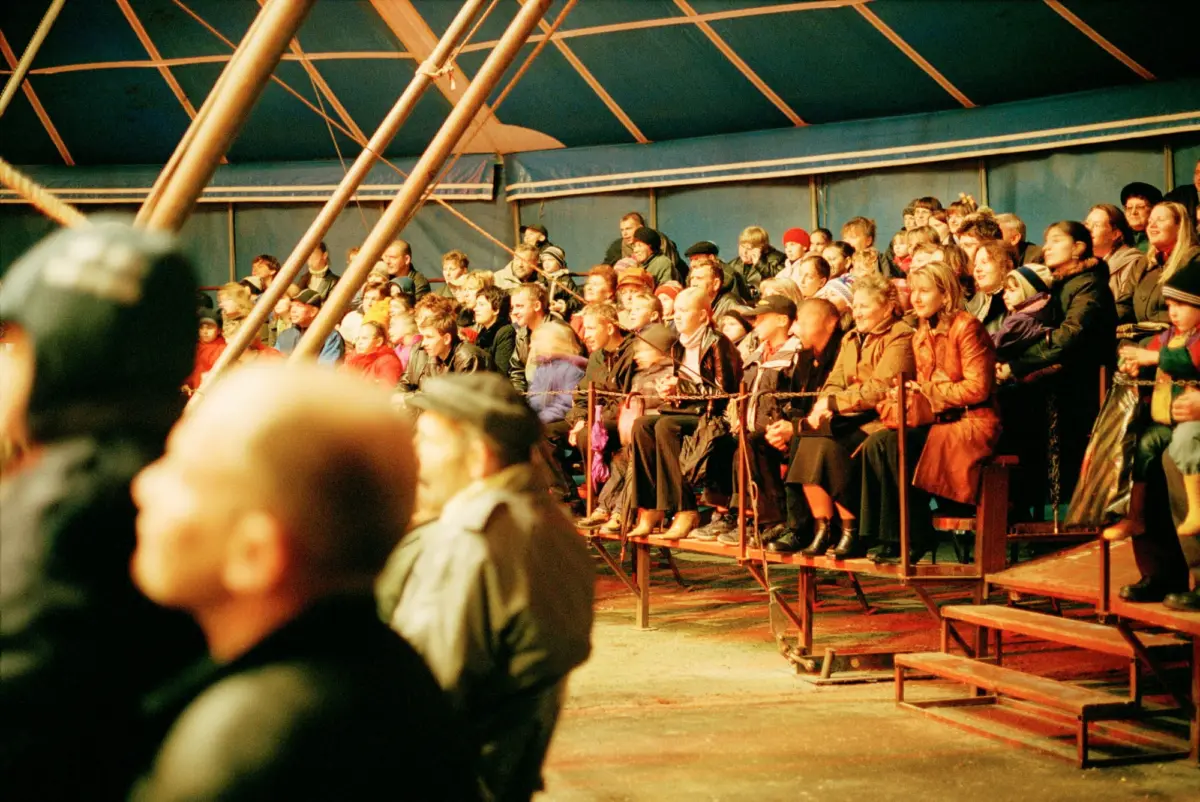 Crowd of spectators seated on bleachers inside a blue striped tent, watching a performance.
