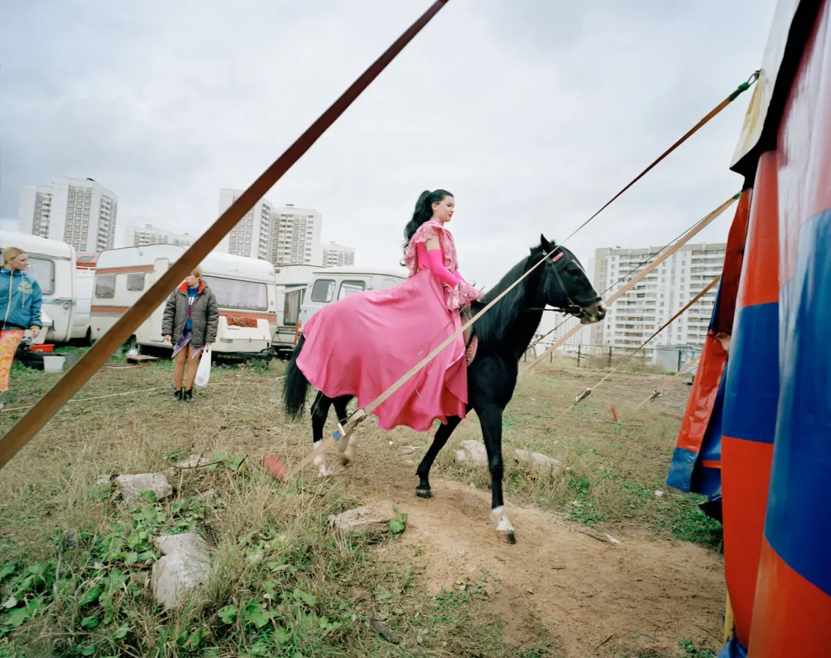 Woman in pink gown rides a black horse at a fairground with caravans and a colorful tent.