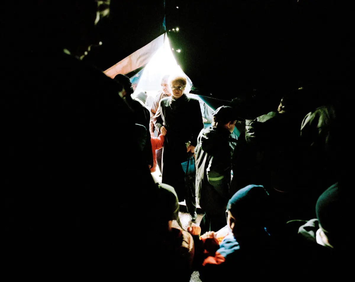 Group of campers standing at the bright open tent doorway in a nighttime campsite.