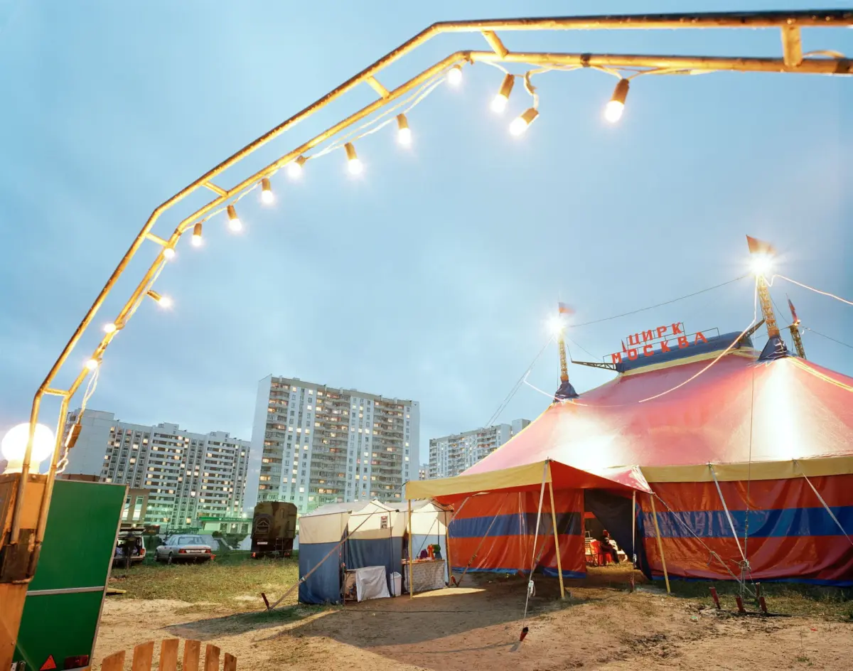 Circus tent with red and blue stripes under string lights at a fairground near apartment buildings.