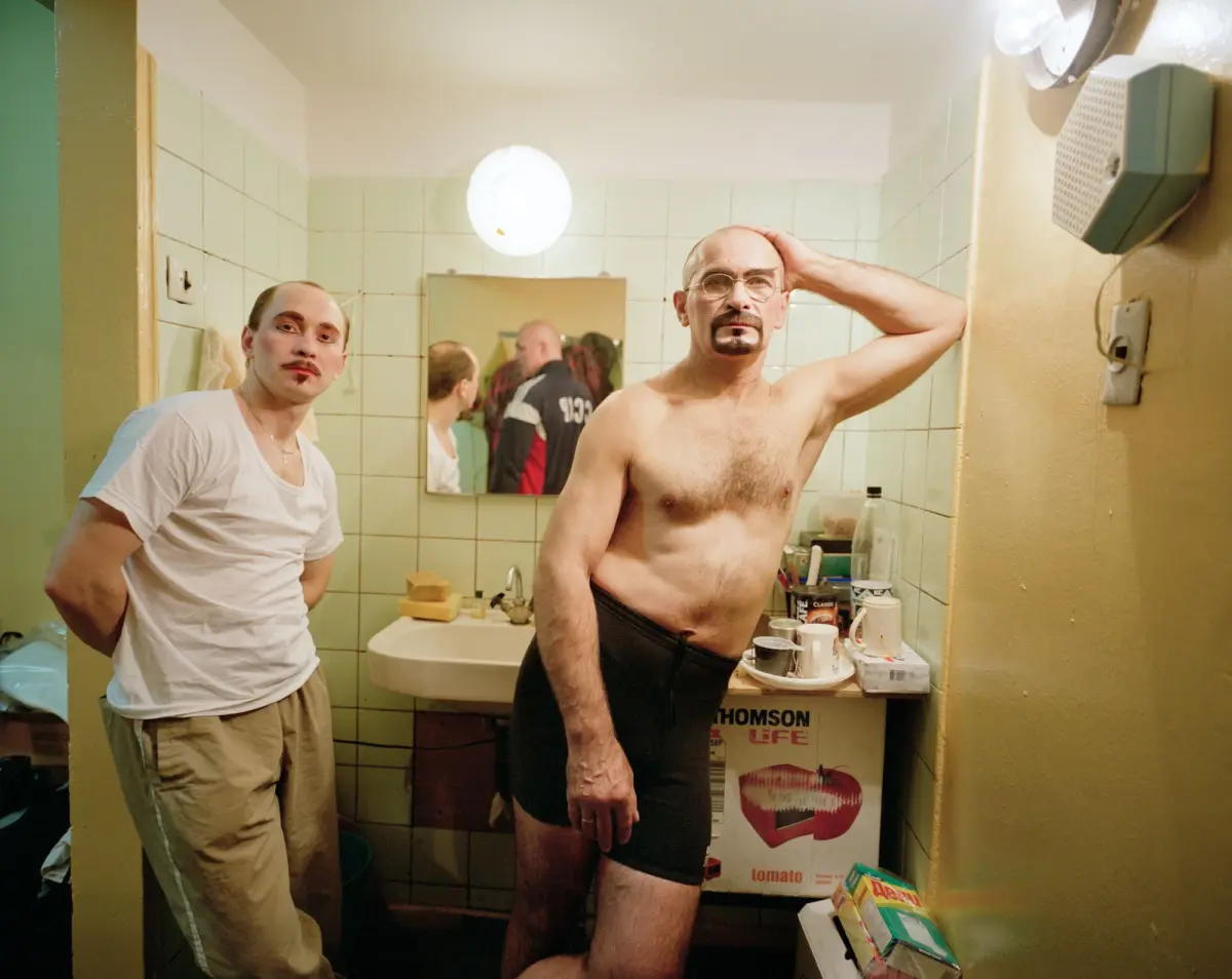 Two adult men in a small tiled bathroom; one shirtless in black shorts, the other in a white T-shirt by the sink with a hand on his head.