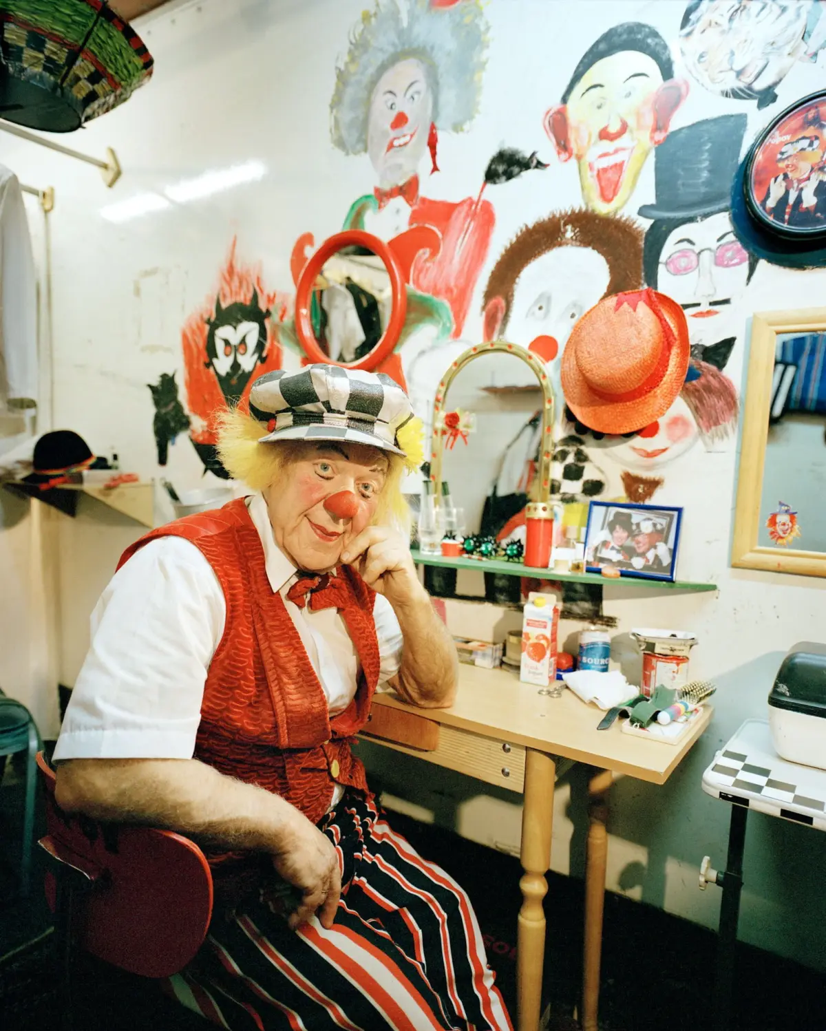 Elderly clown with red nose and yellow hair, wearing striped pants and red vest, sits at a dressing table in a colorful clown-themed room.