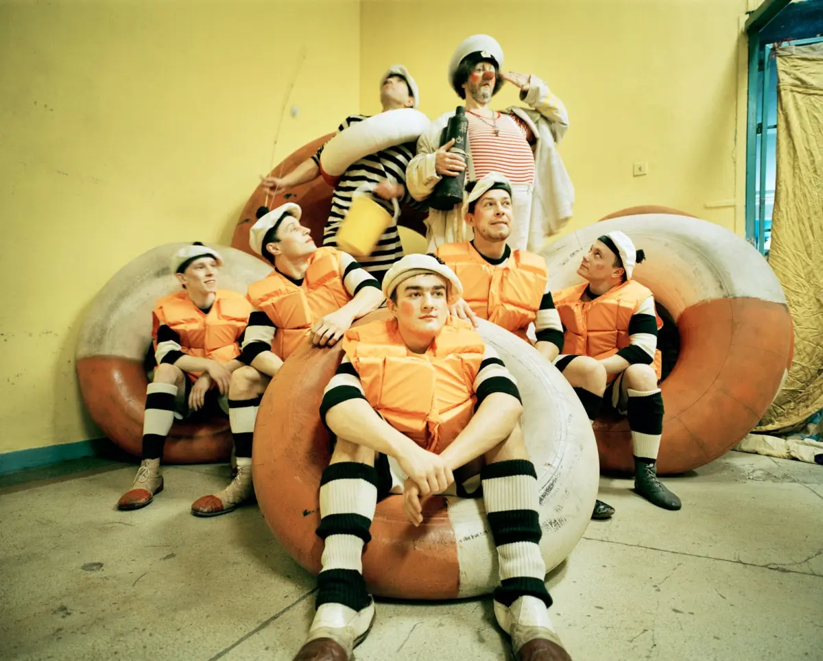 Group of adults wearing orange life jackets pose on large inflatable tubes in a yellow-walled indoor room.