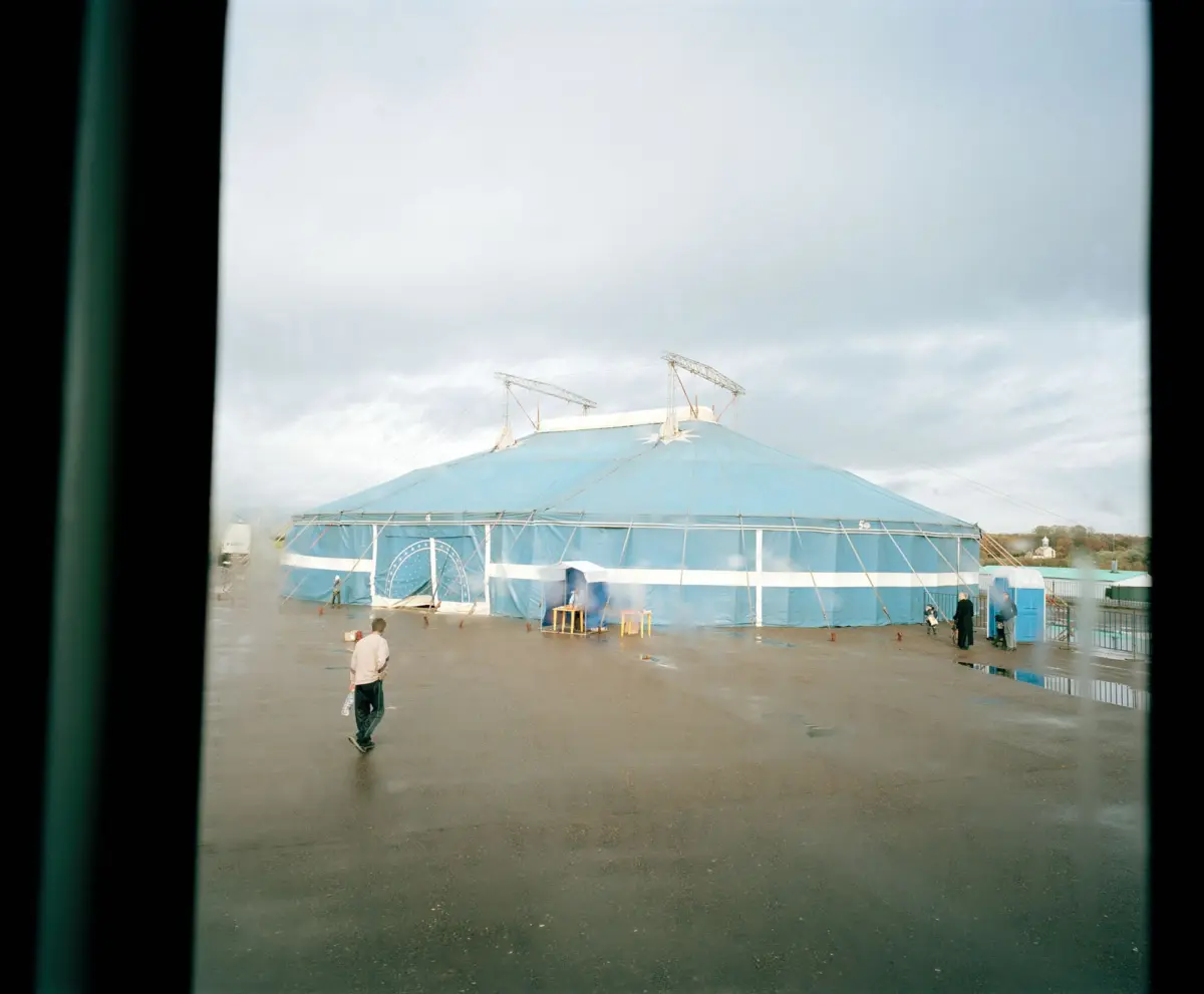 Circular blue circus tent with white stripes on a wet tarmac; several people walk nearby under a gray, overcast sky.