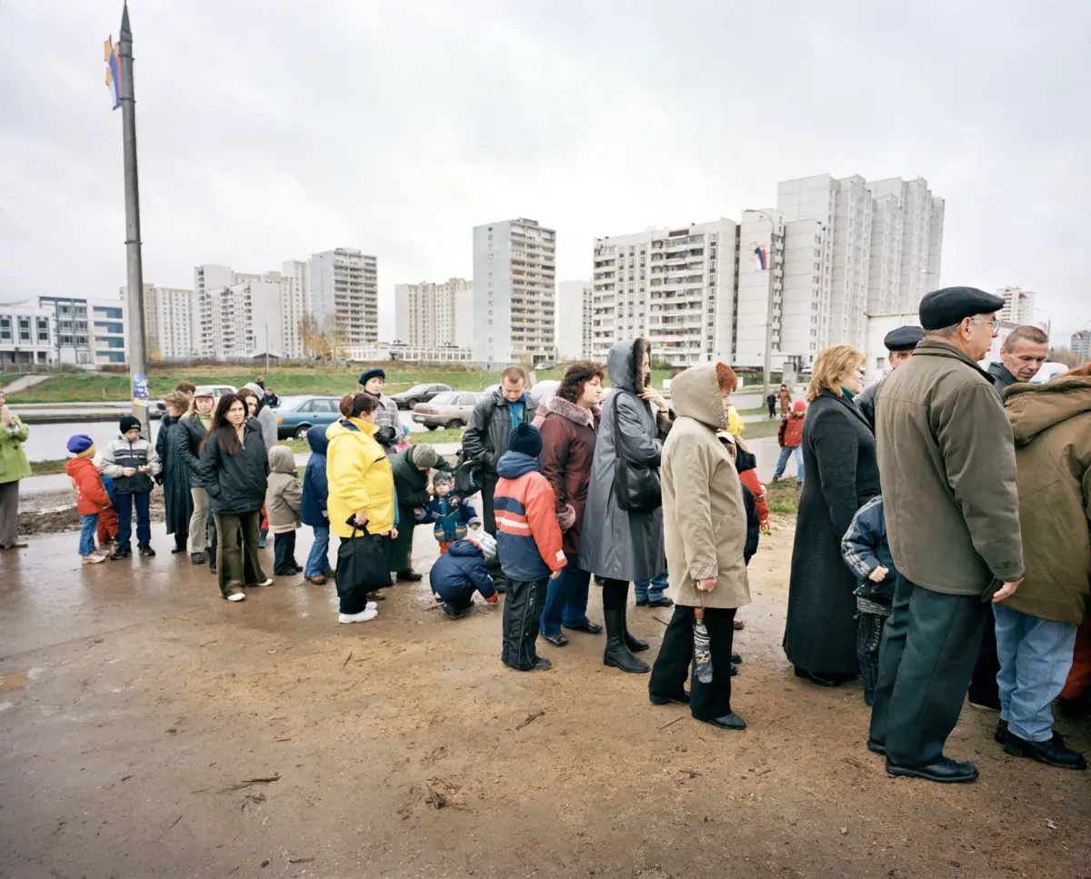 Group of pedestrians in coats and hats walking along a muddy path beside a river, with high-rise apartment blocks in the background.