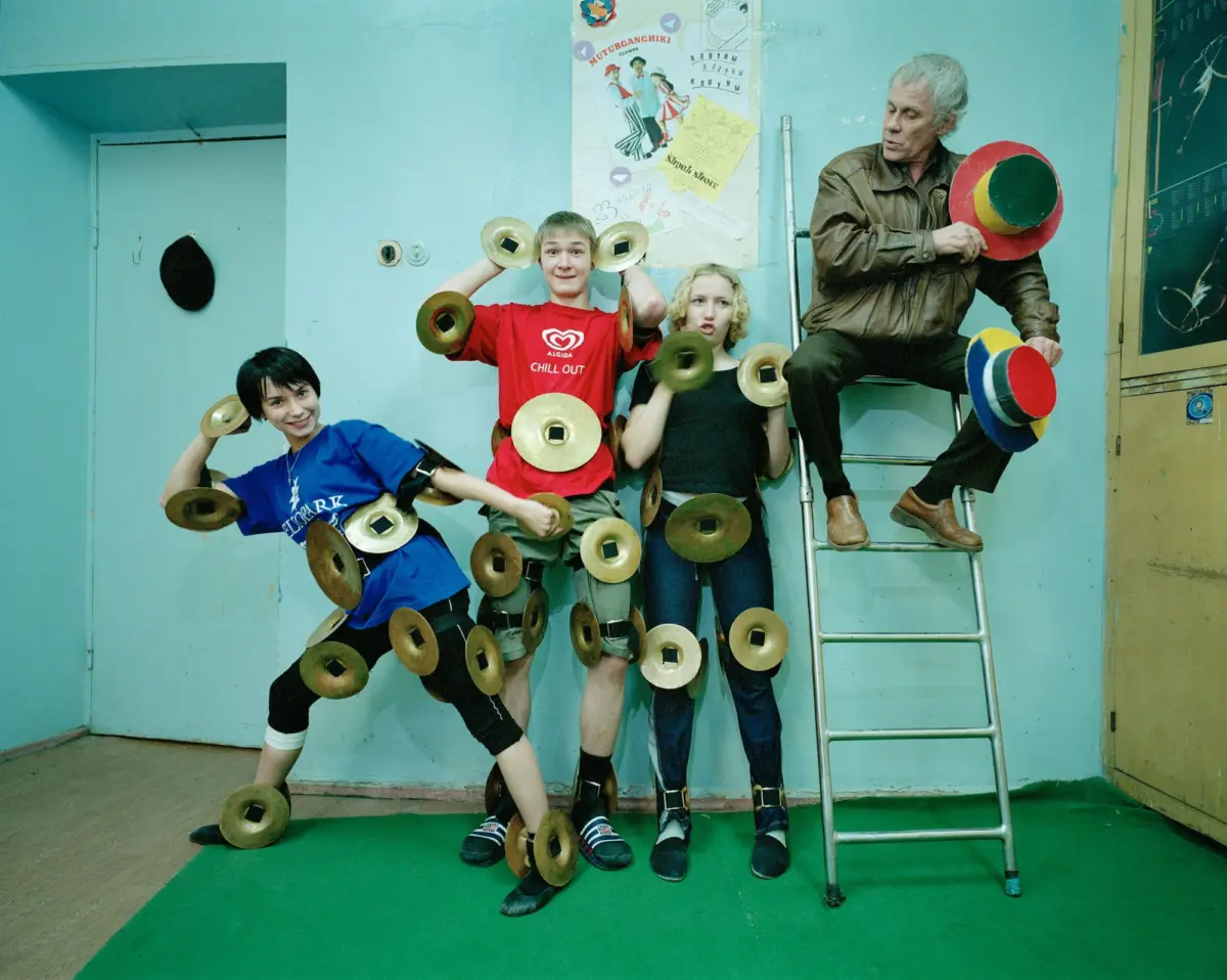 Five people posing indoors; children wearing cymbals strapped to their bodies, adult on a ladder holding colorful hats.
