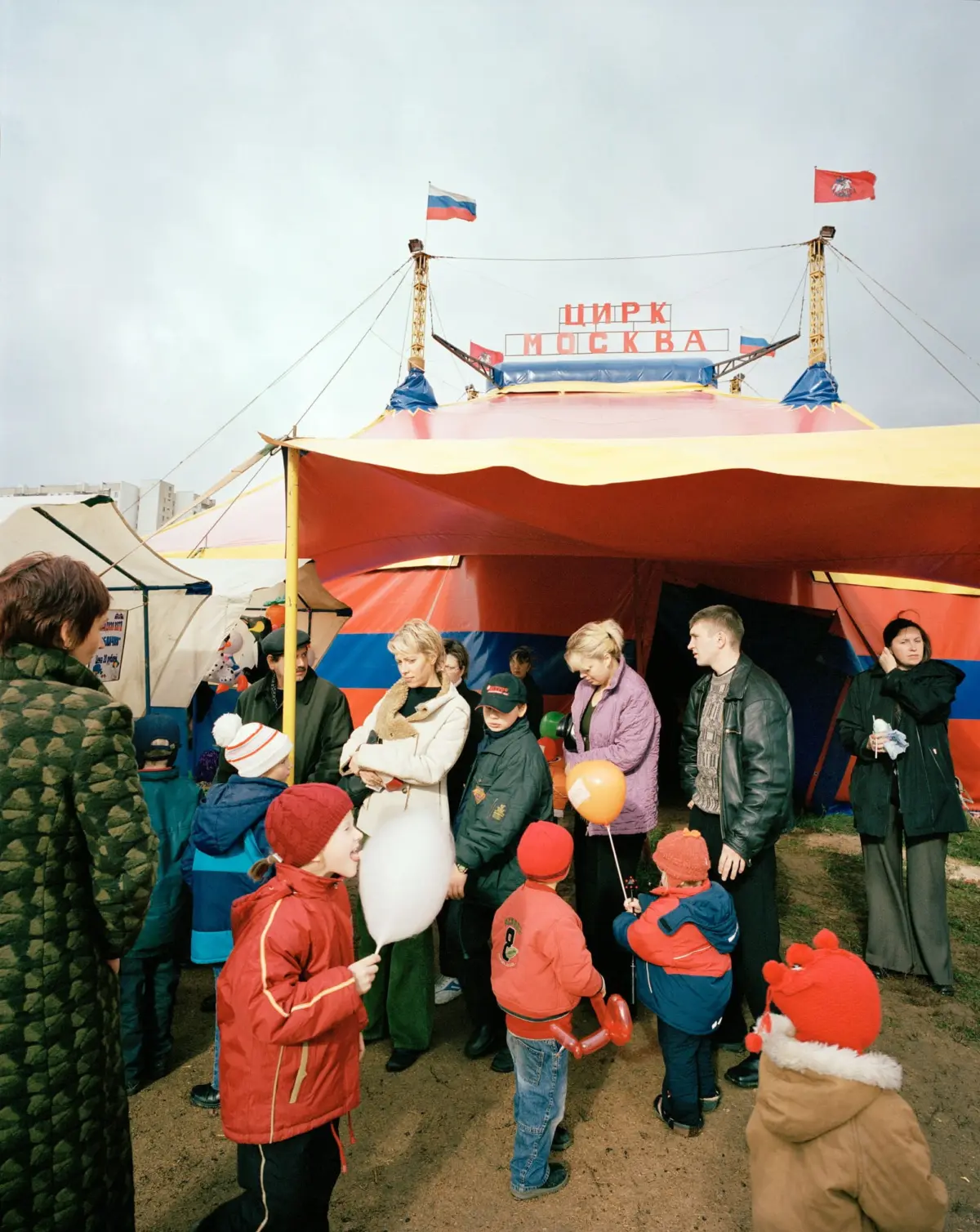 Crowd of adults and children gathers around a colorful circus tent; Circus Moscow sign ЦИРК МОСКВА with Russian flags atop towers.