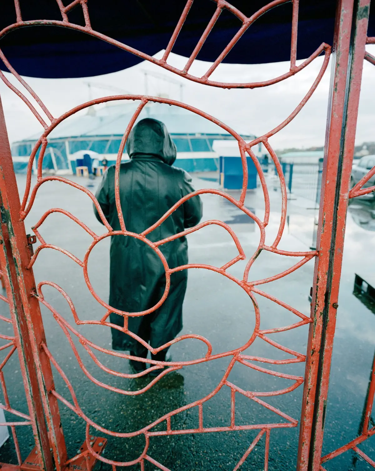 Hooded figure in a dark raincoat stands behind a red floral wrought-iron gate on a wet waterfront promenade.