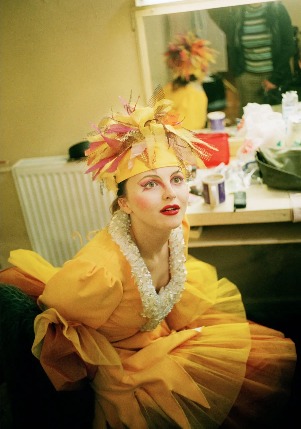 Performer in yellow-orange theatrical gown with elaborate feather headpiece and white lace collar, seated in a cluttered dressing room backstage.