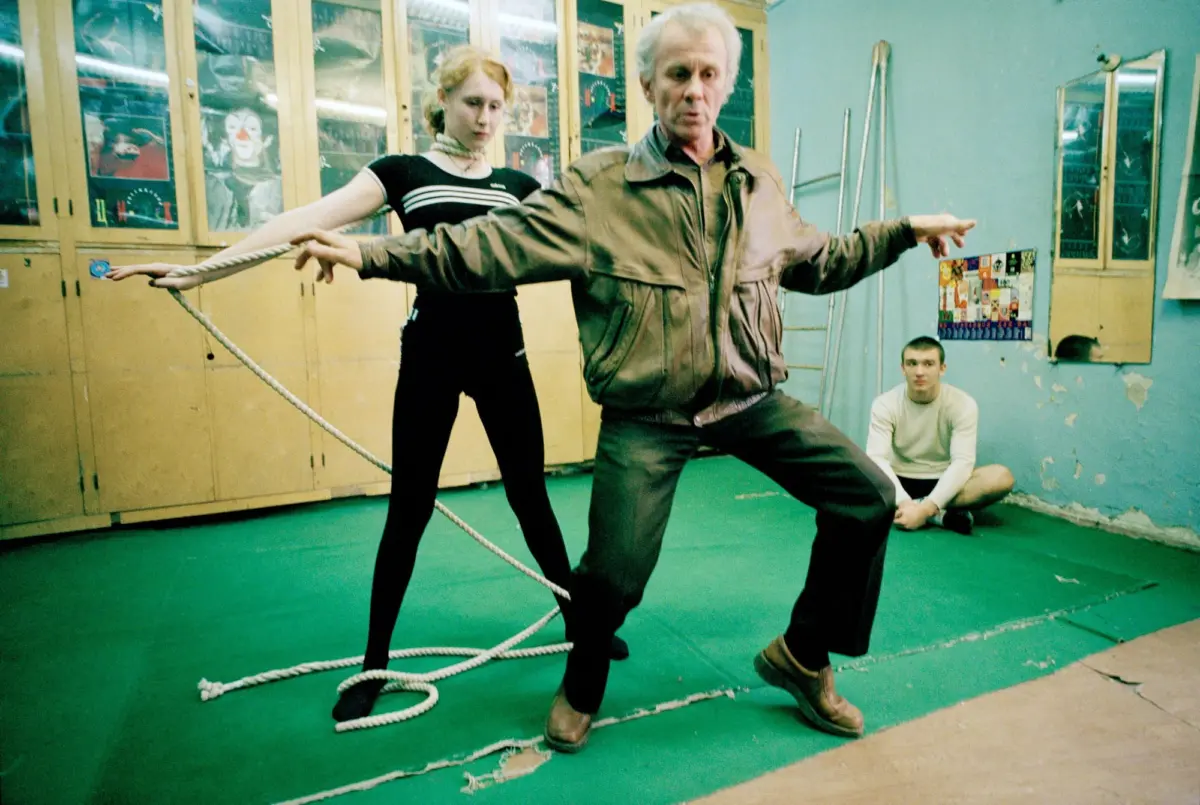 Older adult in brown leather jacket balances on rope in a gym-like room; younger adult assists; green floor, wooden lockers, spectator seated.