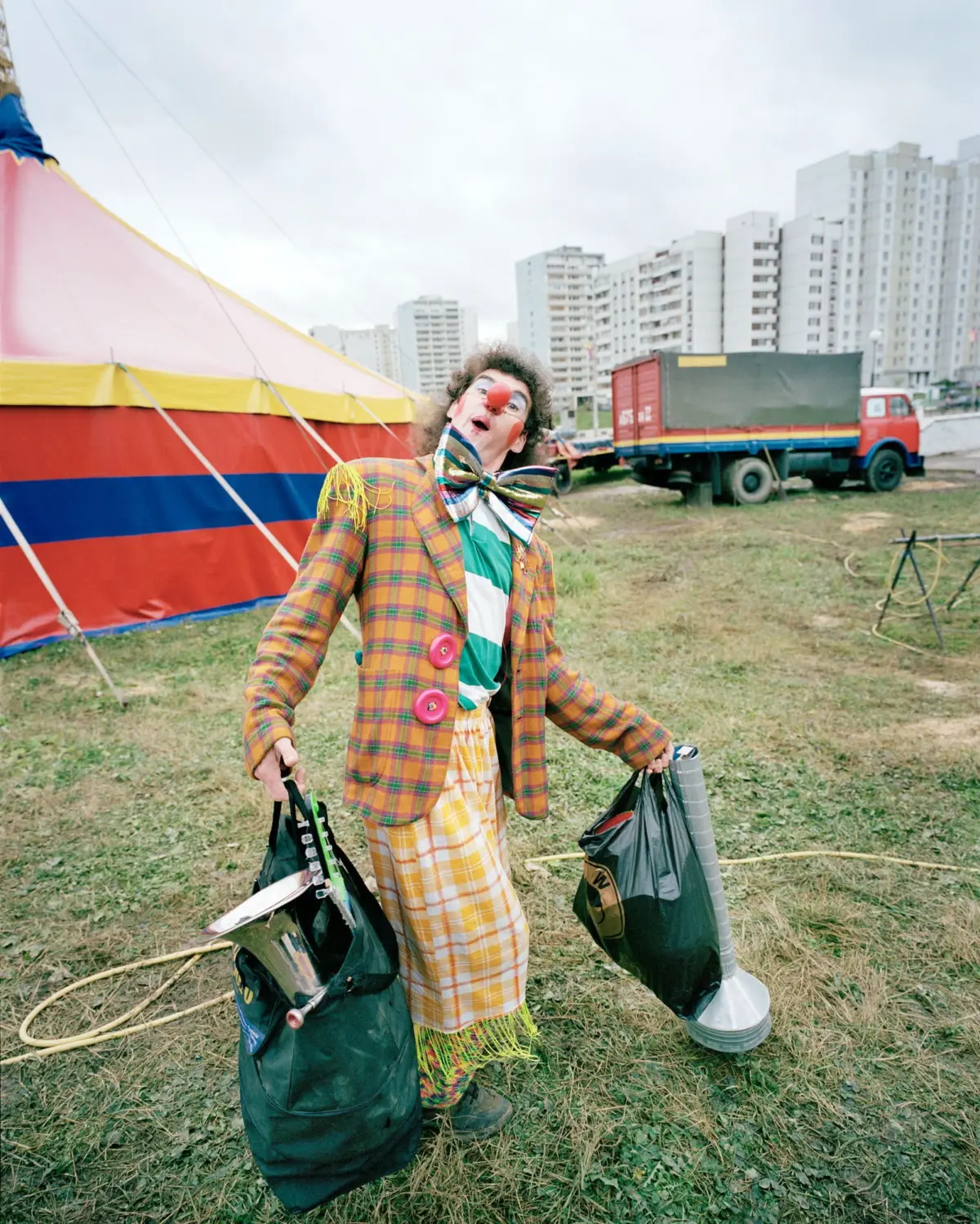 Clown in plaid suit and oversized bow tie, carrying two prop bags beside a colorful circus tent, with urban high-rise buildings in the background.