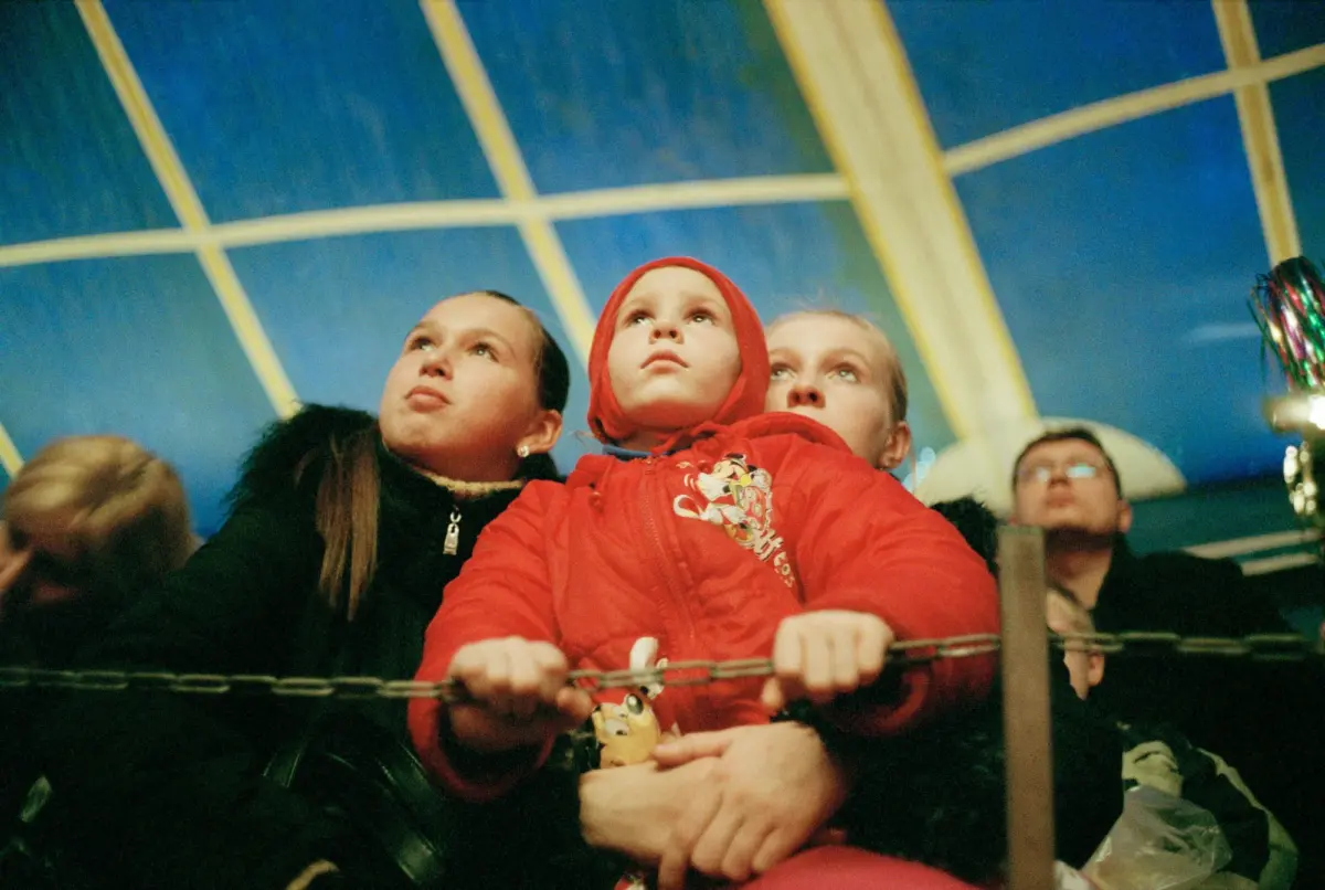 Child in red hooded jacket gazes upward, gripping a railing on an indoor amusement ride.