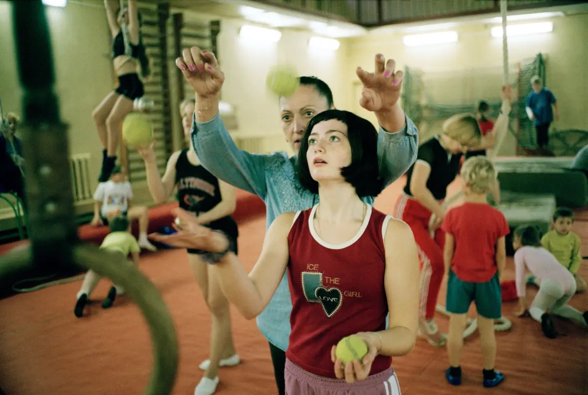 Woman with short black hair in a red tank top holds a yellow ball as a trainer guides her in a crowded gymnasium.