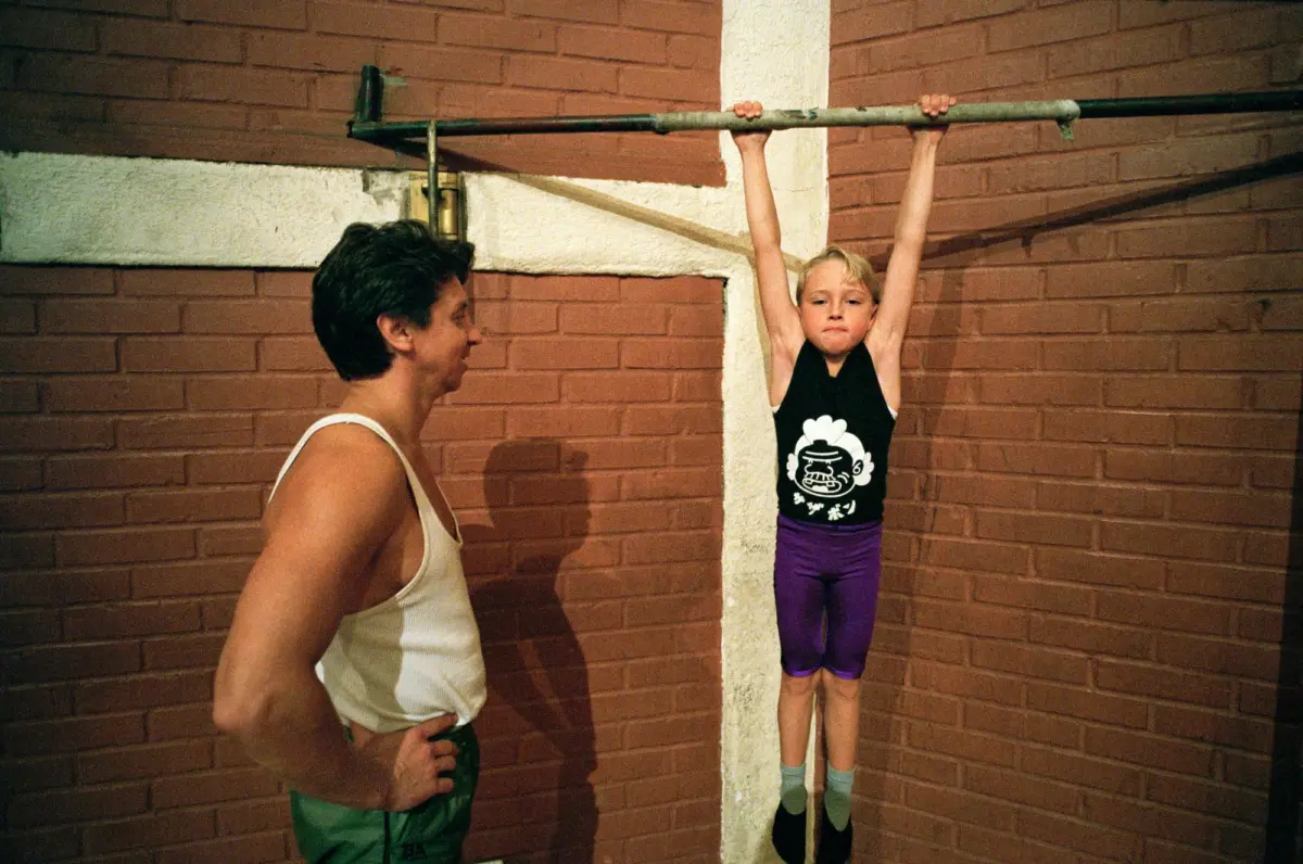 Child hangs from a horizontal bar with arms raised against a white pillar between red brick walls; adult man stands nearby.