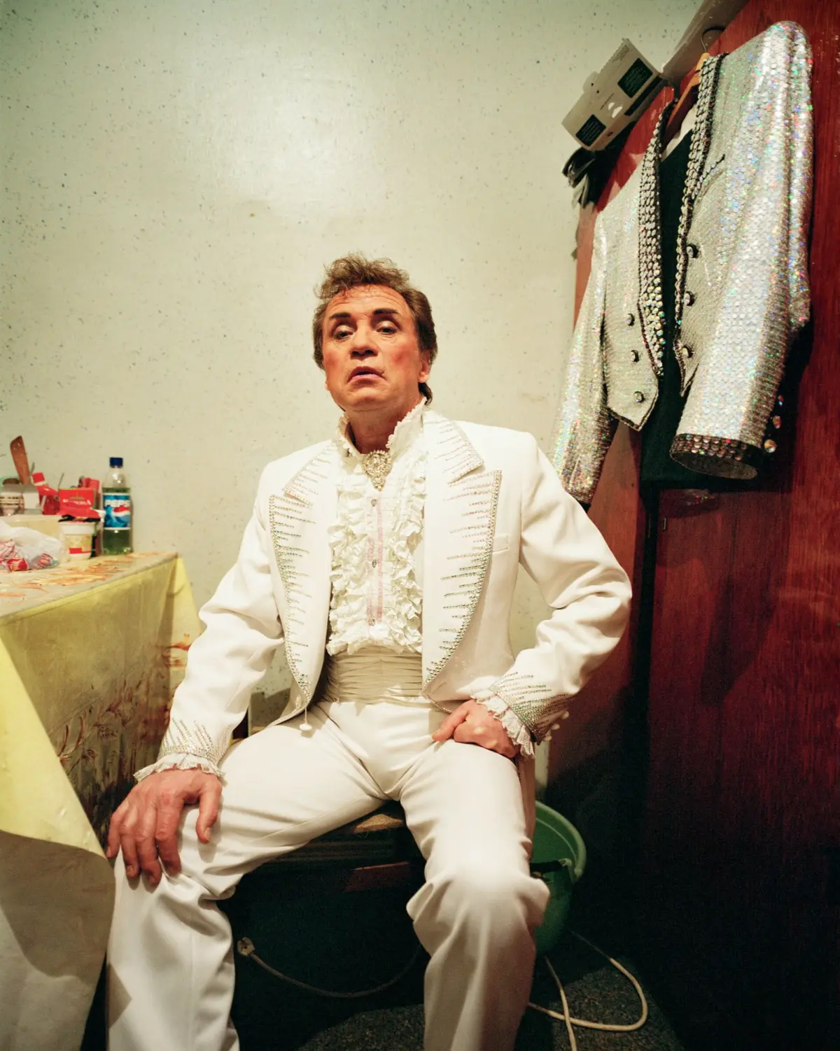 Elvis-style performer in a white sequined suit sits in a backstage dressing room, with jackets hanging nearby.