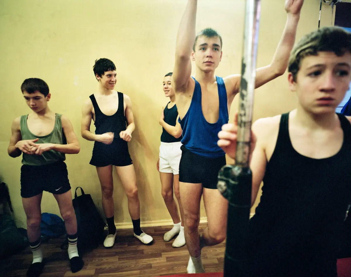 Group of teenage boys in athletic tank tops and shorts in a gym; one raises arms beside a metal pole while peers watch.