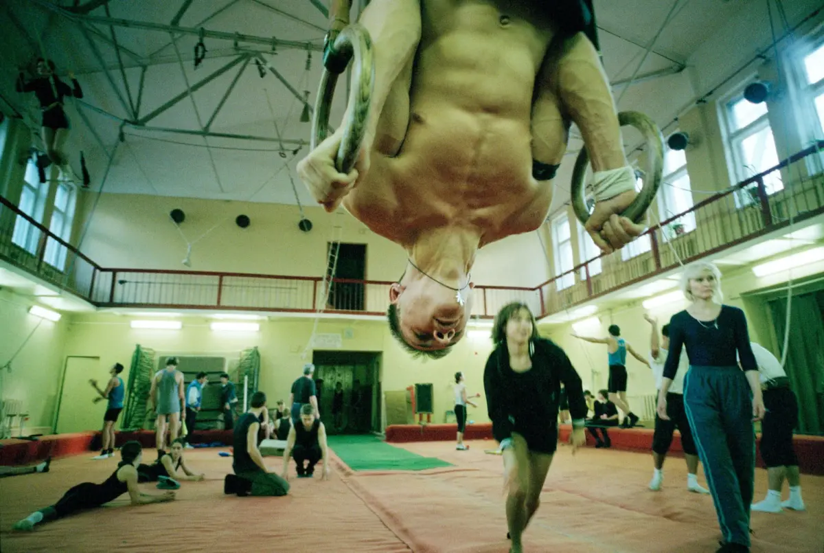 Person hanging upside down from gymnastic rings in a gymnasium with mats and other participants nearby.