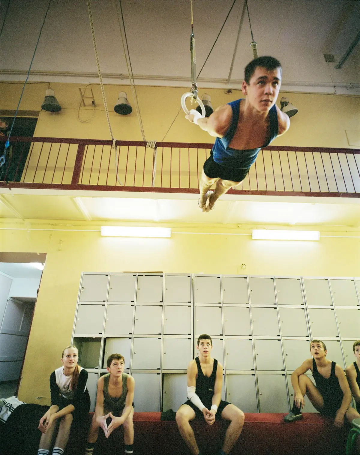 Gymnast in blue singlet swinging on gymnastic rings indoors, spectators seated by a locker wall.