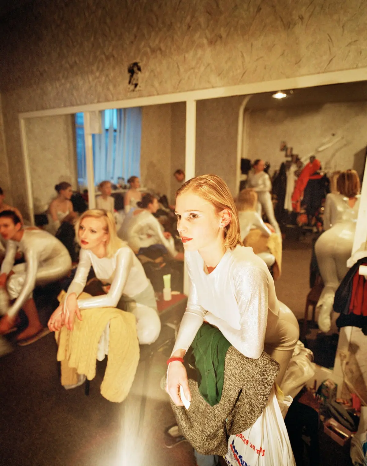 Backstage dressing room with models in metallic outfits; foreground model sits on a bench in a white shimmering top, leaning forward.