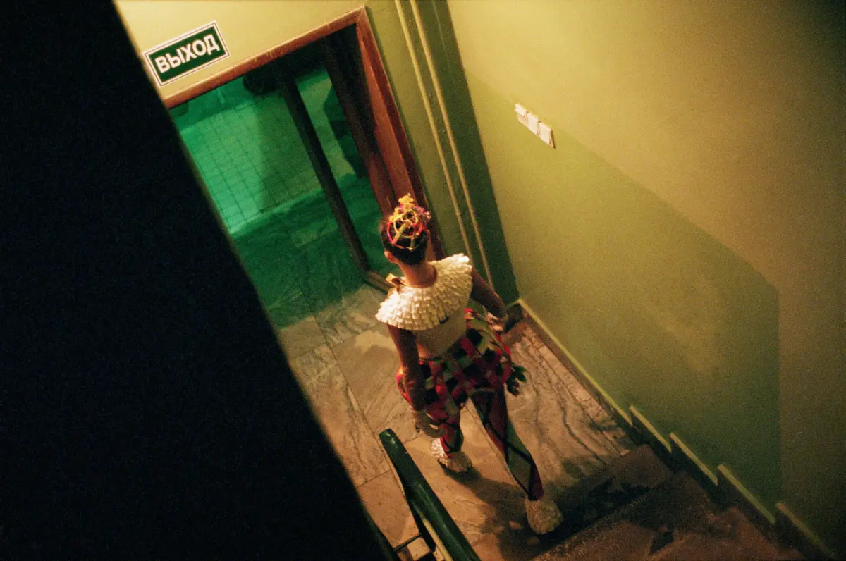 Performer in a colorful headpiece and large white ruffled collar, descending a green-walled indoor stairwell.