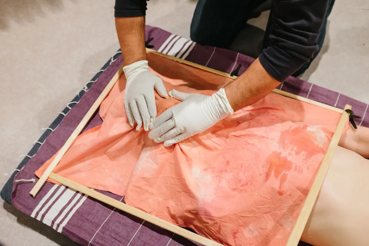 Gloved hands smooth orange fabric inside a wooden coffin frame atop a purple striped blanket.