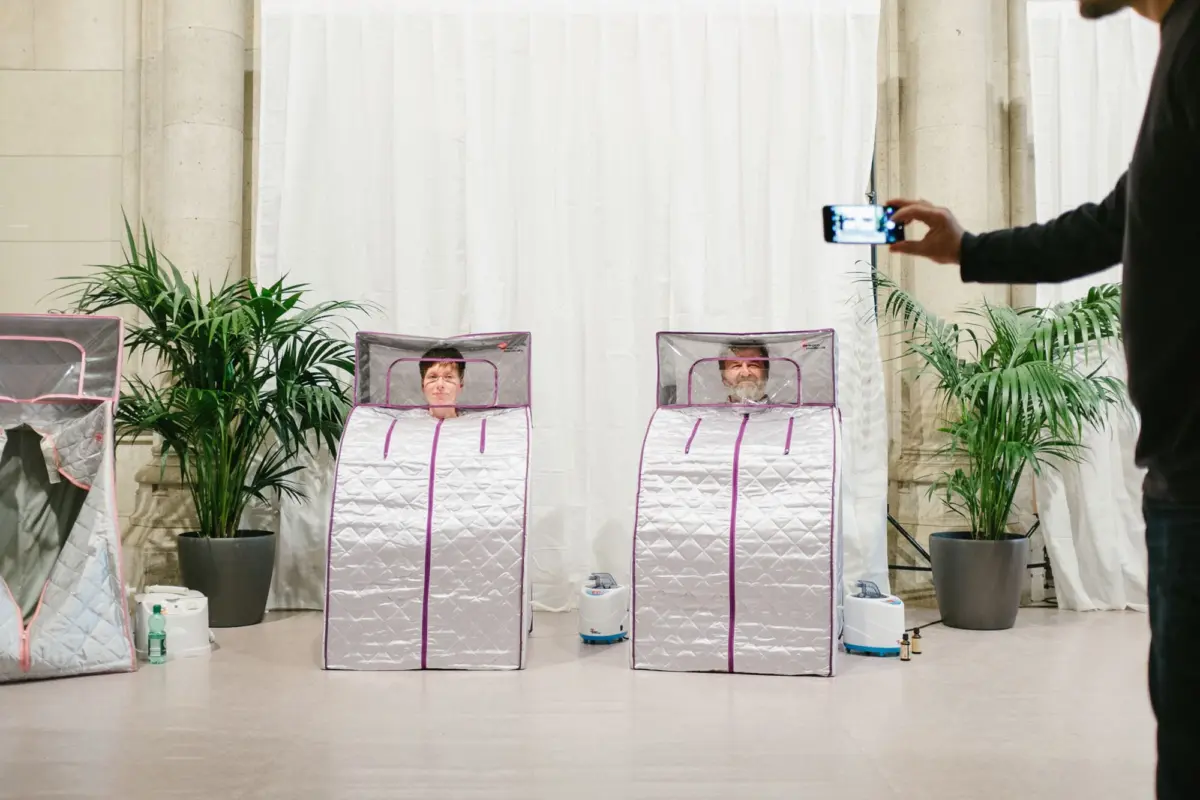 Two people sit inside silver quilted sleeping pods; a person films them with a smartphone in a bright studio with potted plants and white curtains.