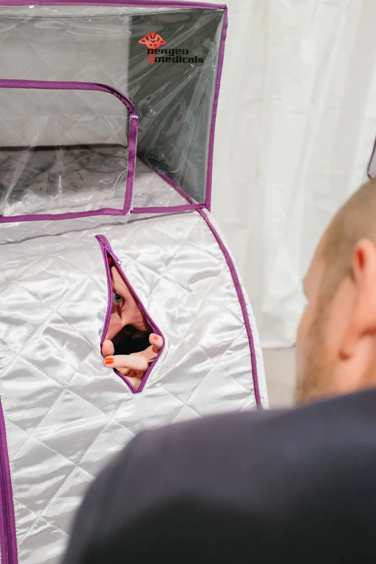 Person peering through a diamond-quilted silver suit with purple trim; cut-out window near chest, Newgen Medicals logo visible on the suit panel.