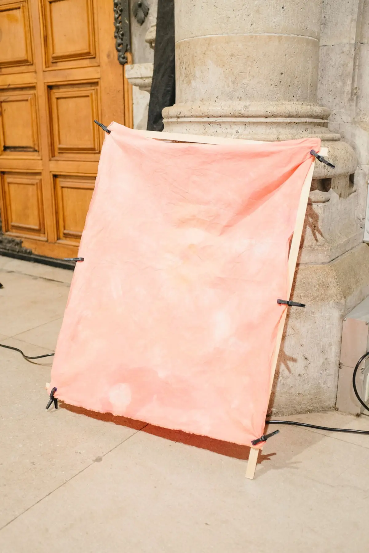 Peach-colored fabric screen attached to a wooden frame with clips, set on a sidewalk beside a stone column and wooden door.
