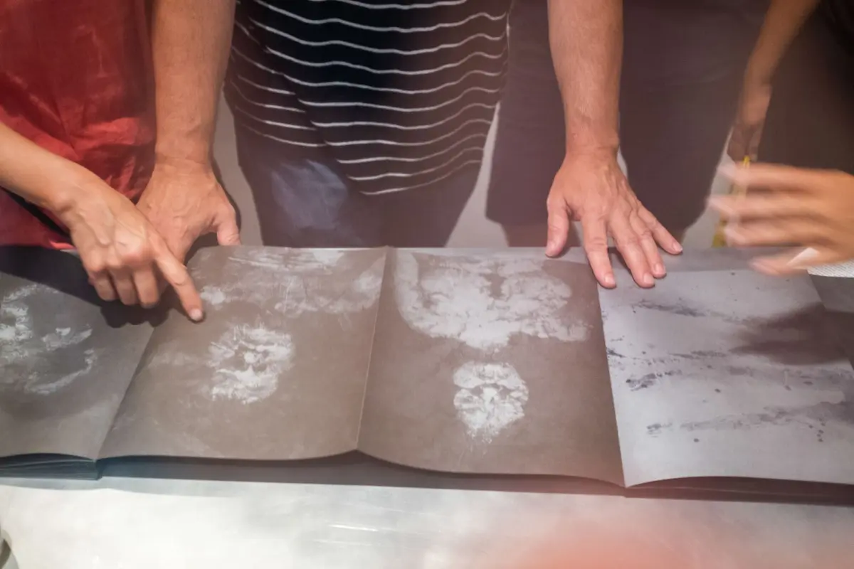 Three adults press hands on a large open dusty art book on a workshop table.