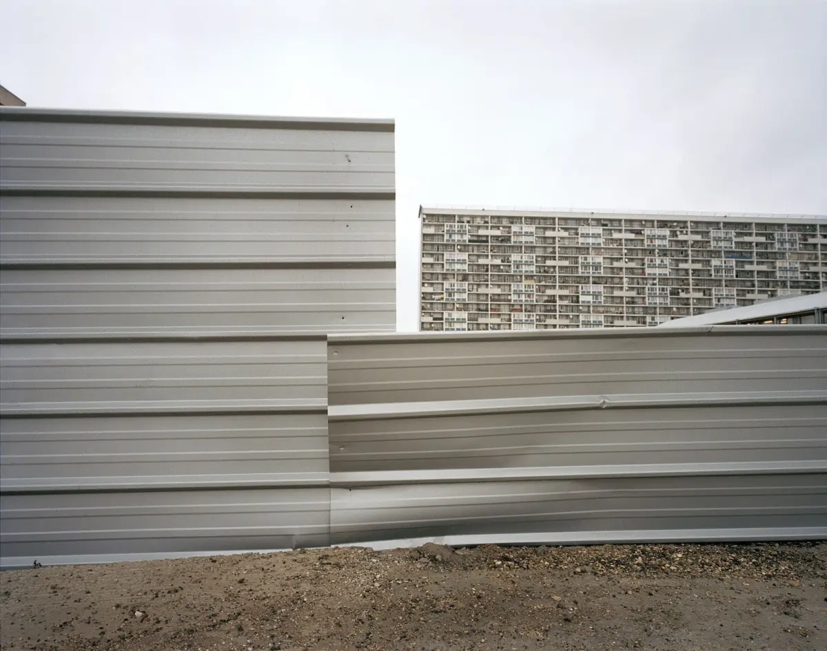 Construction site barrier panels in the foreground; dirt ground and a multi-story apartment block in the background beneath a pale sky.