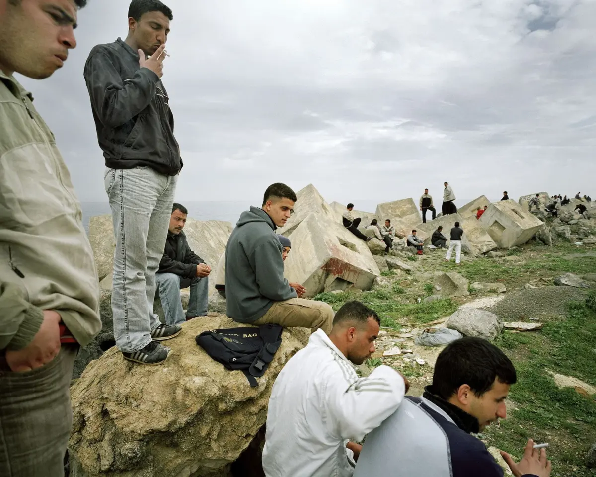 Group of men on a rocky shoreline, some sitting on large concrete blocks and others standing, with a cloudy sky and sea in the distance.