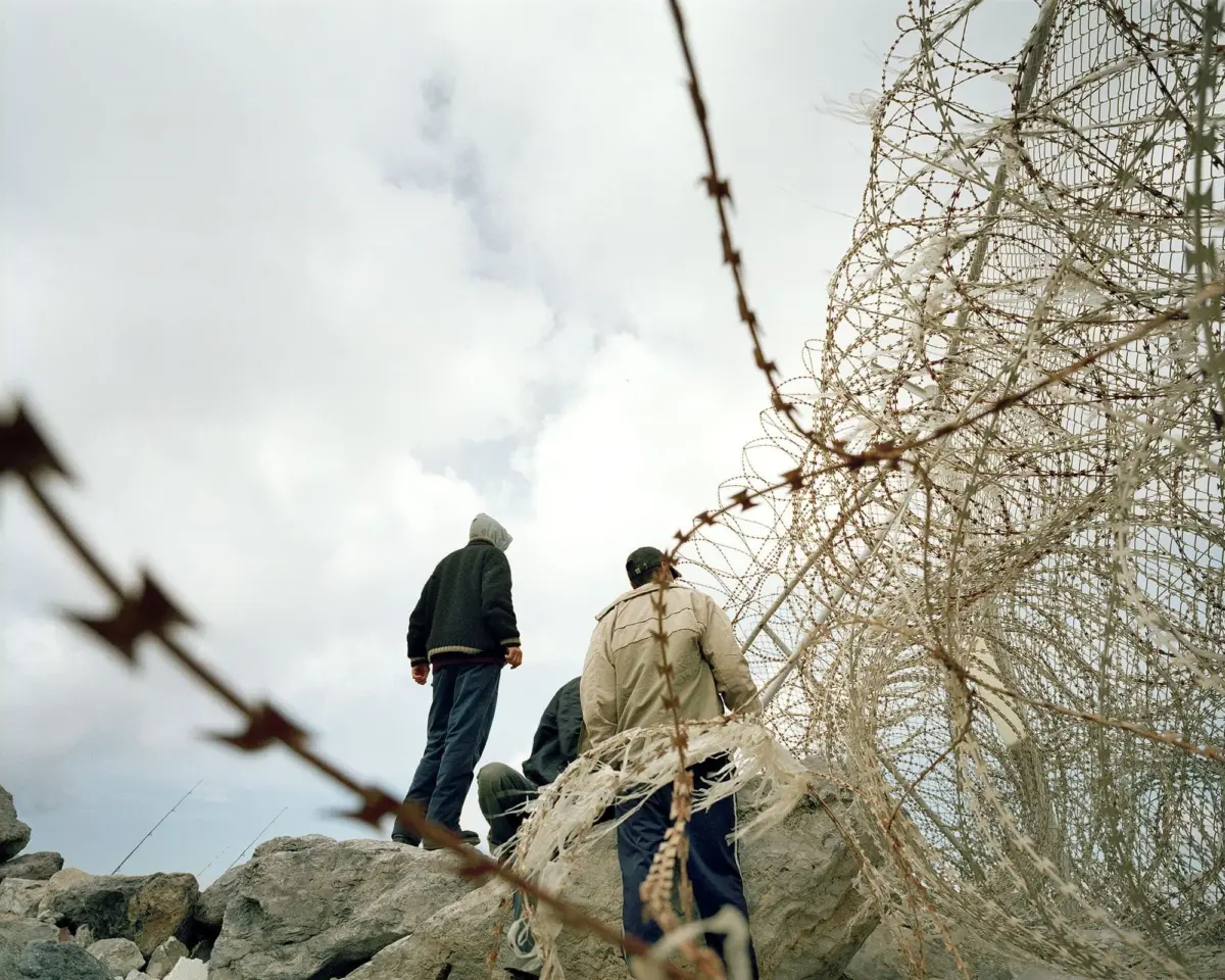 Three people stand on jagged rocks beside a tall chain-link fence topped with razor wire under a cloudy sky.