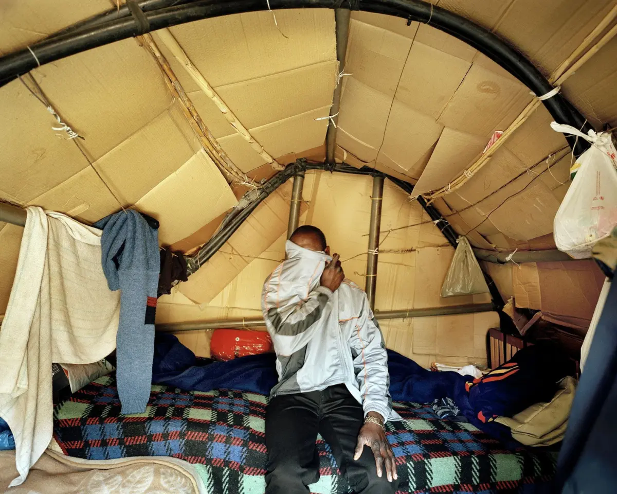 Person wearing a light gray jacket and face mask stands inside a circular cardboard shelter with exposed pipes and blankets around.