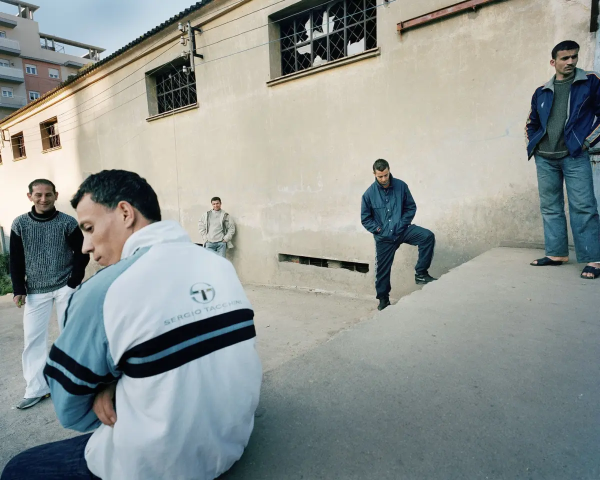 Person wearing light blue jacket sits with arms crossed in the foreground; four others stand along a beige building with barred windows.