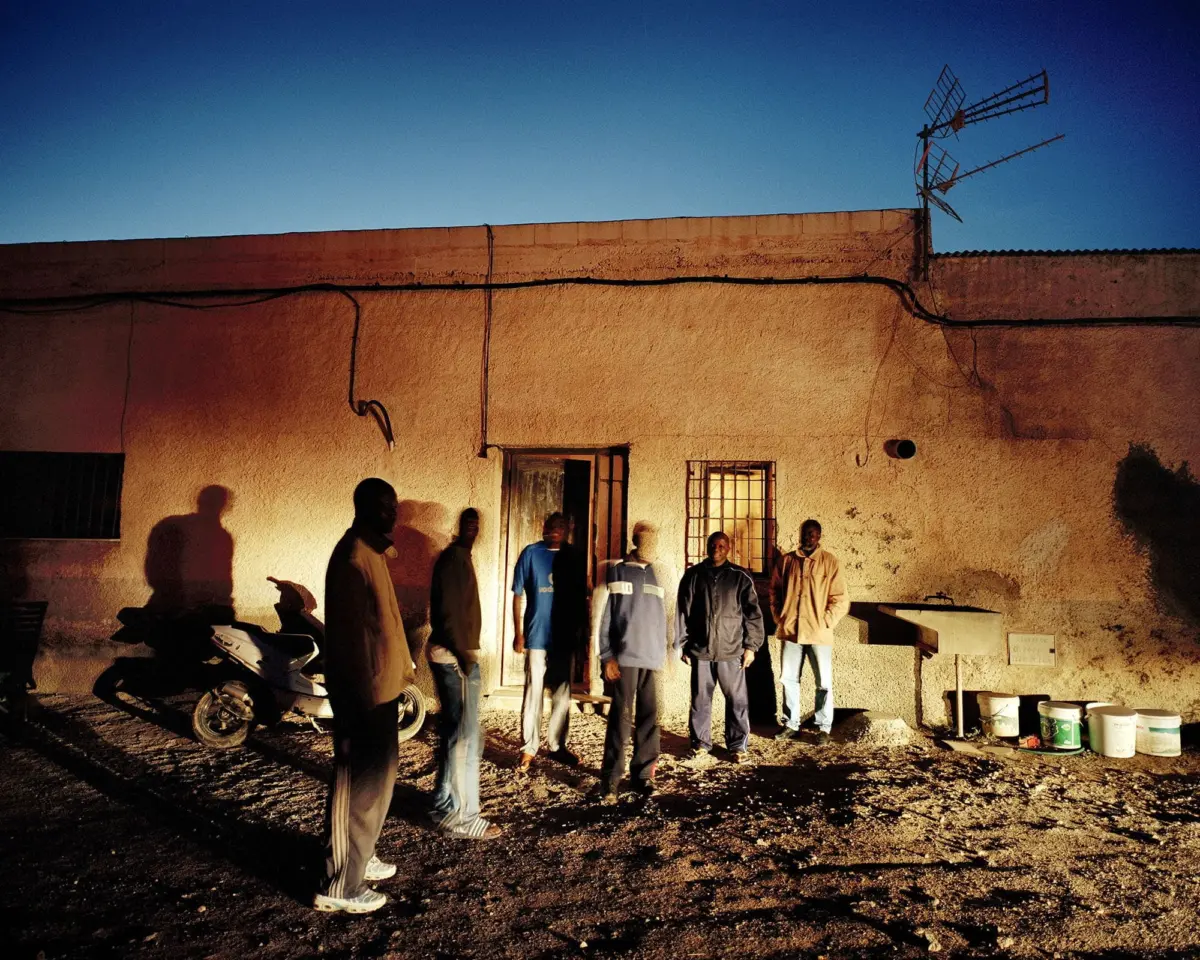 Group of men standing in front of a beige stucco building at dusk, with exposed cables, a scooter, and a rooftop antenna against a blue sky.