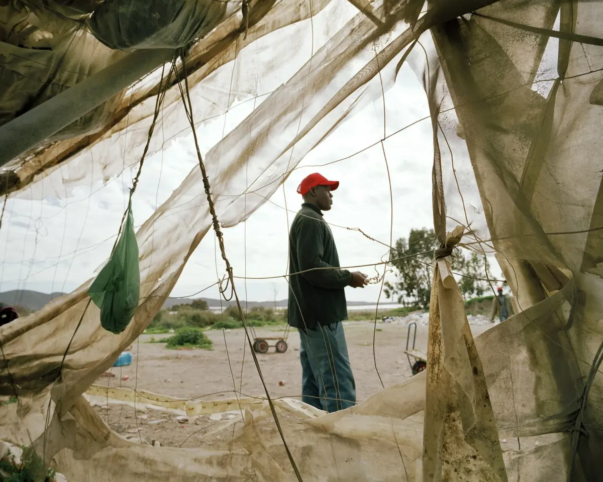 Person wearing a red cap stands inside a torn canvas tent, adjusting a rope in a barren, dusty field.