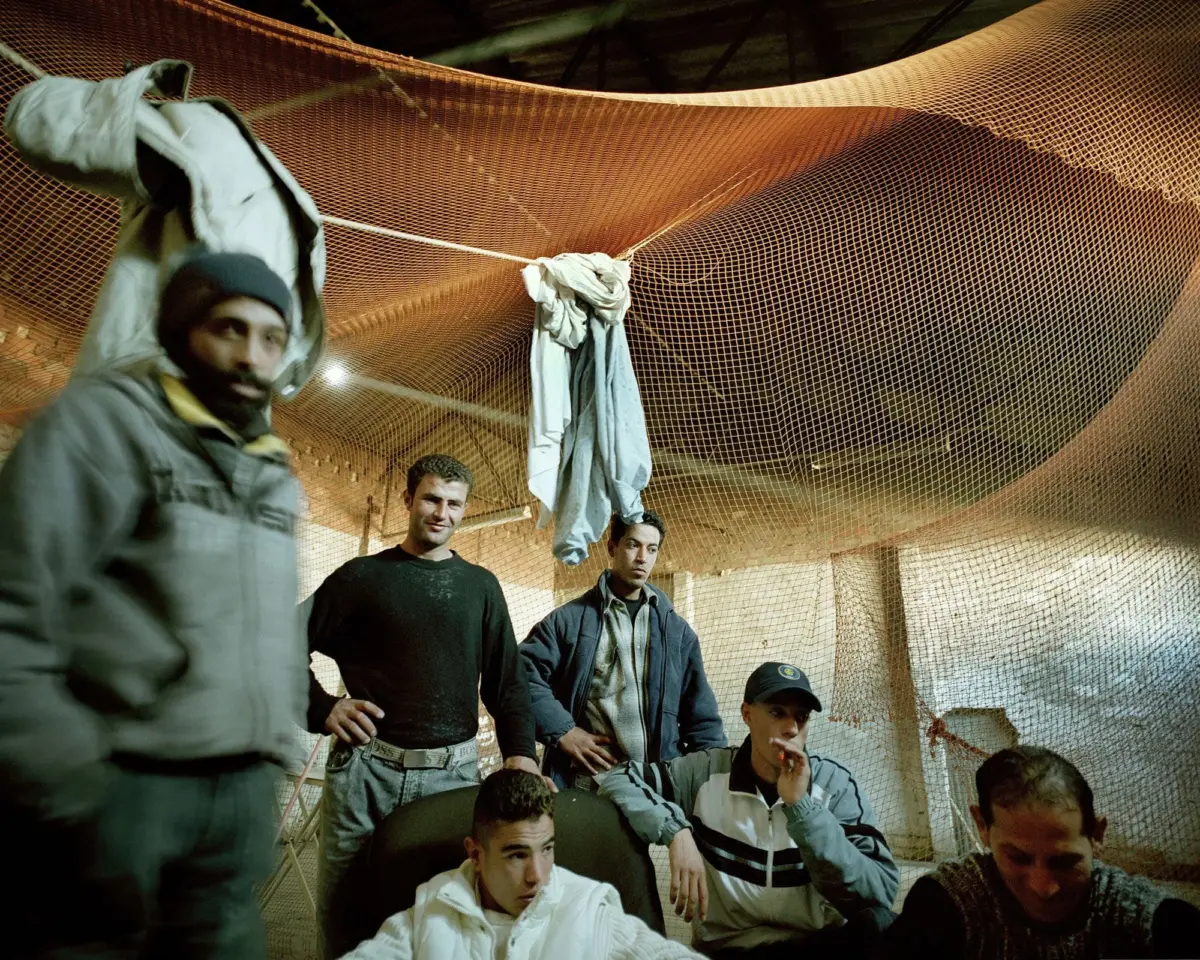 Group of men in an industrial workshop examining a large orange mesh net hanging from ropes.