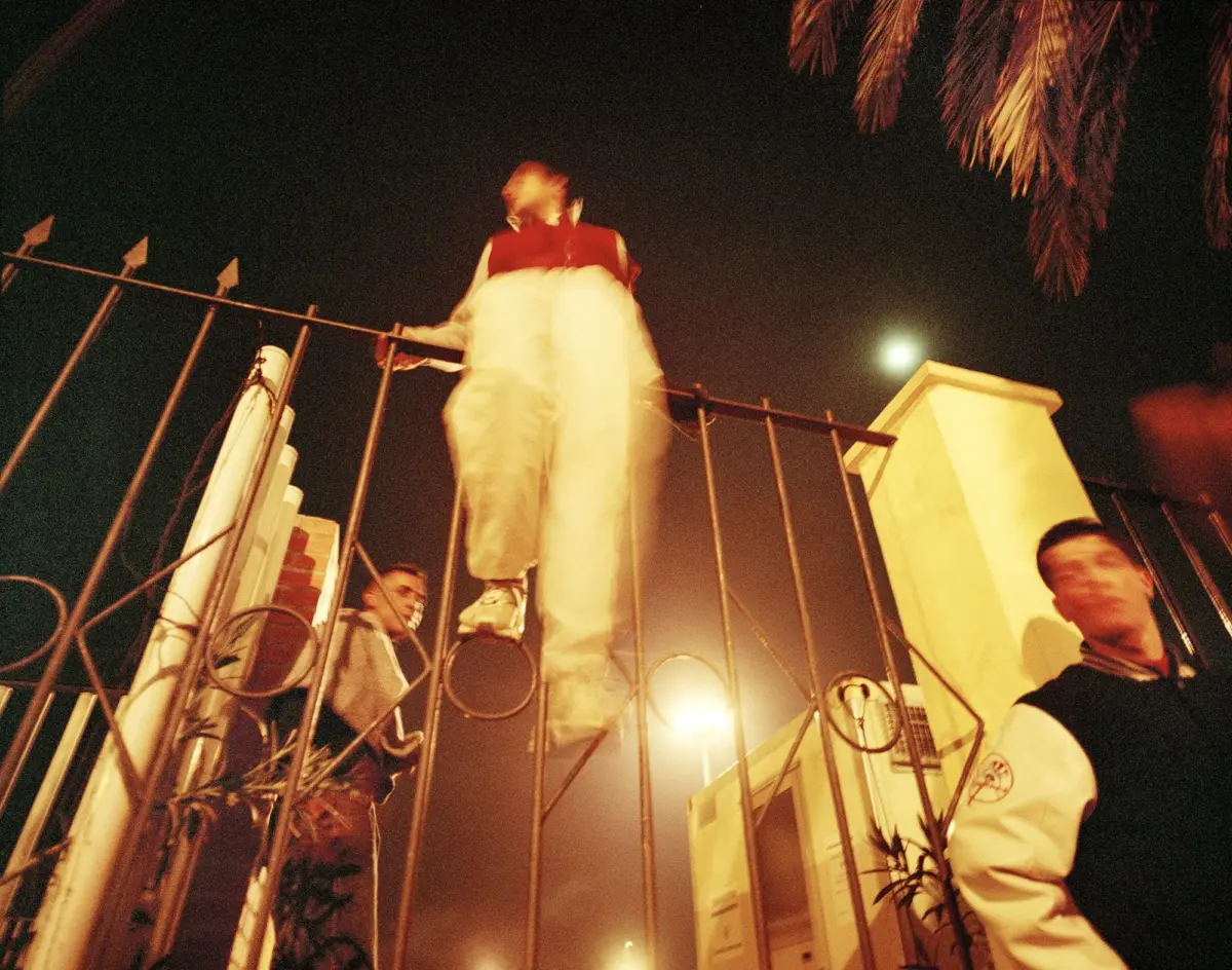 Climber wearing red top and white pants atop a metal gate at night, with security guards nearby and palm trees overhead.
