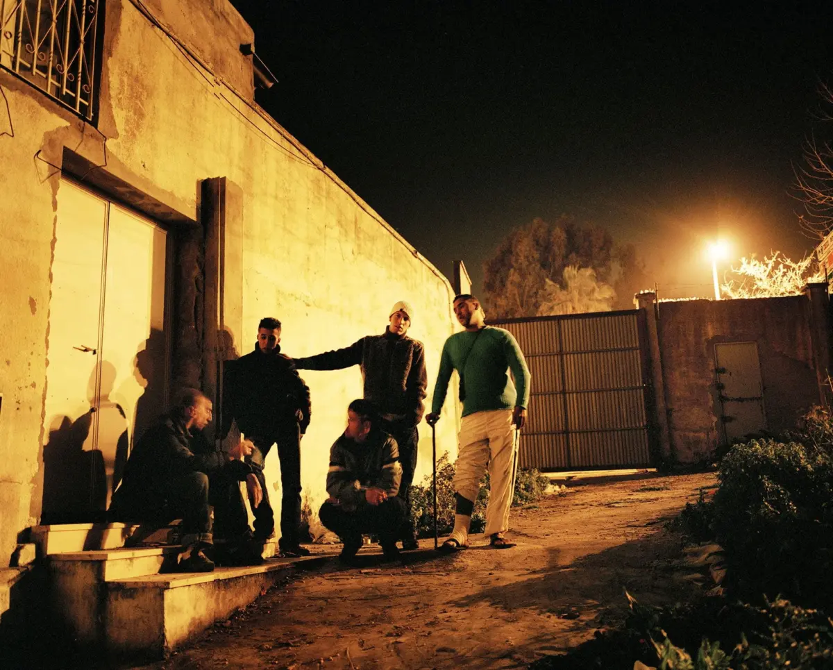 Group of men at night outside a weathered building; some sit on steps while others stand and converse near a gate by orange streetlight.