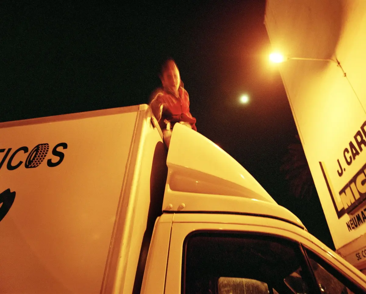 Person standing on top of a white semi-truck cab at night, beside a large trailer under a streetlight.