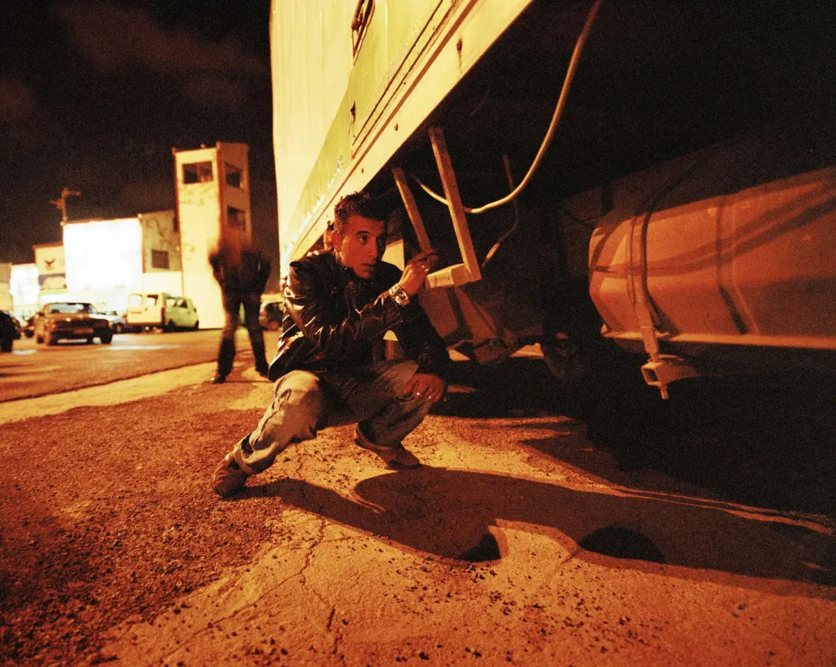 Man in a leather jacket crouches beside a truck trailer frame on a dimly lit urban street at night, inspecting a metal structure.