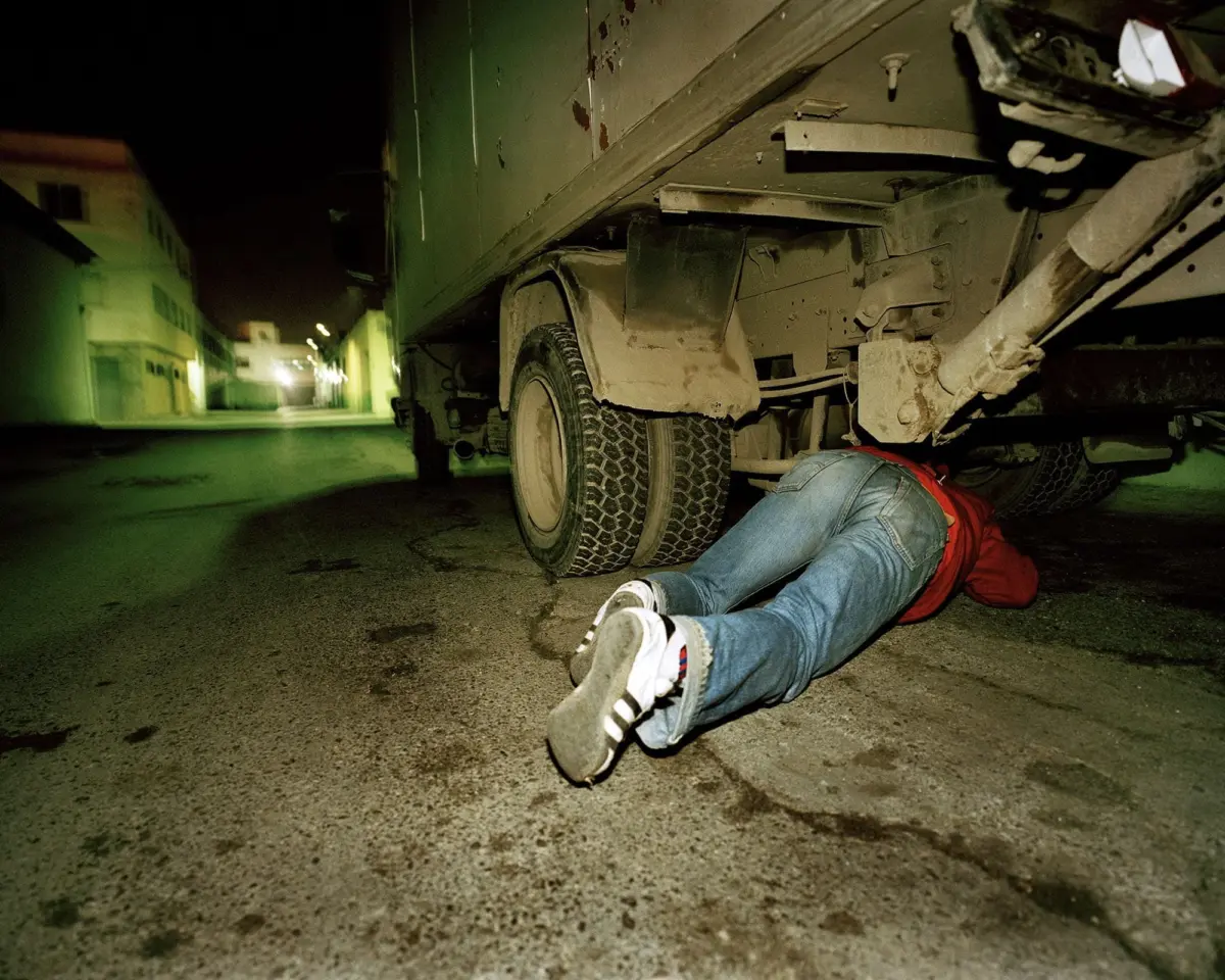 Person lying on their stomach under the rear of a large truck on a dim, green-lit urban street at night.