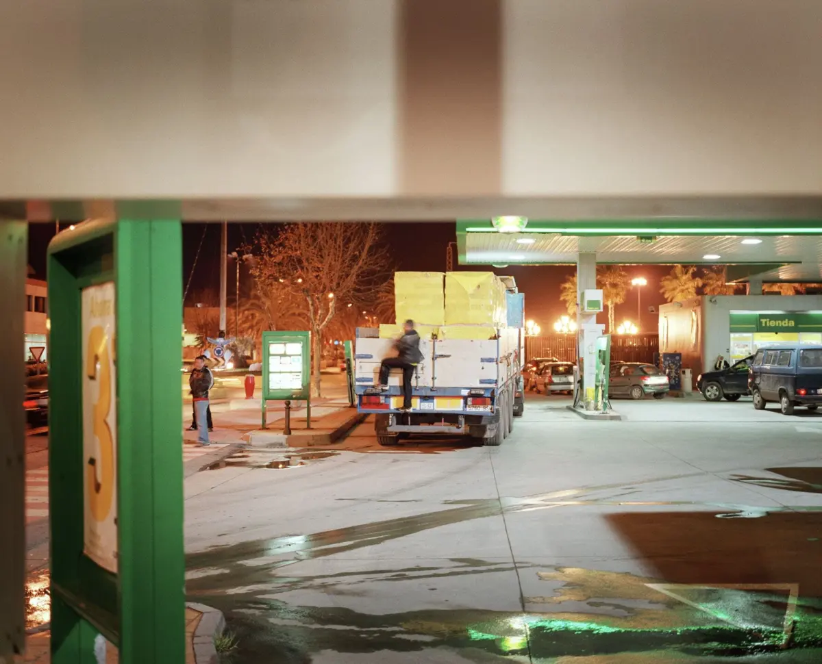 Night-time gas station forecourt; a flatbed truck stacked with boxes, with a worker standing on the truck.