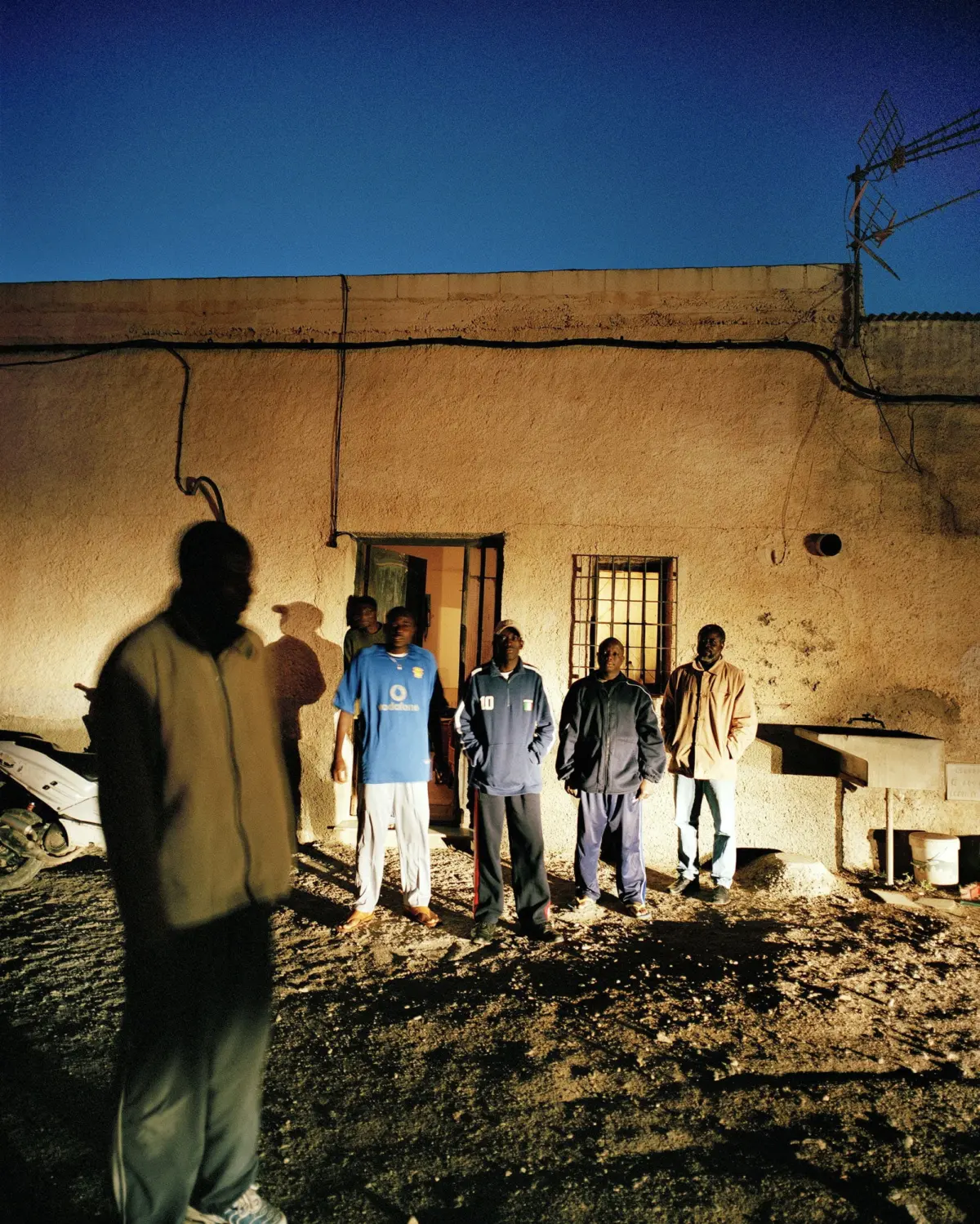 Group of men standing outside a stucco building at night, doorway and barred window, blue sky and overhead wires.