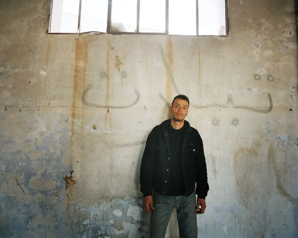 Adult person stands against a weathered graffiti-covered wall in an abandoned building; wearing a dark jacket and jeans, with a barred window above.