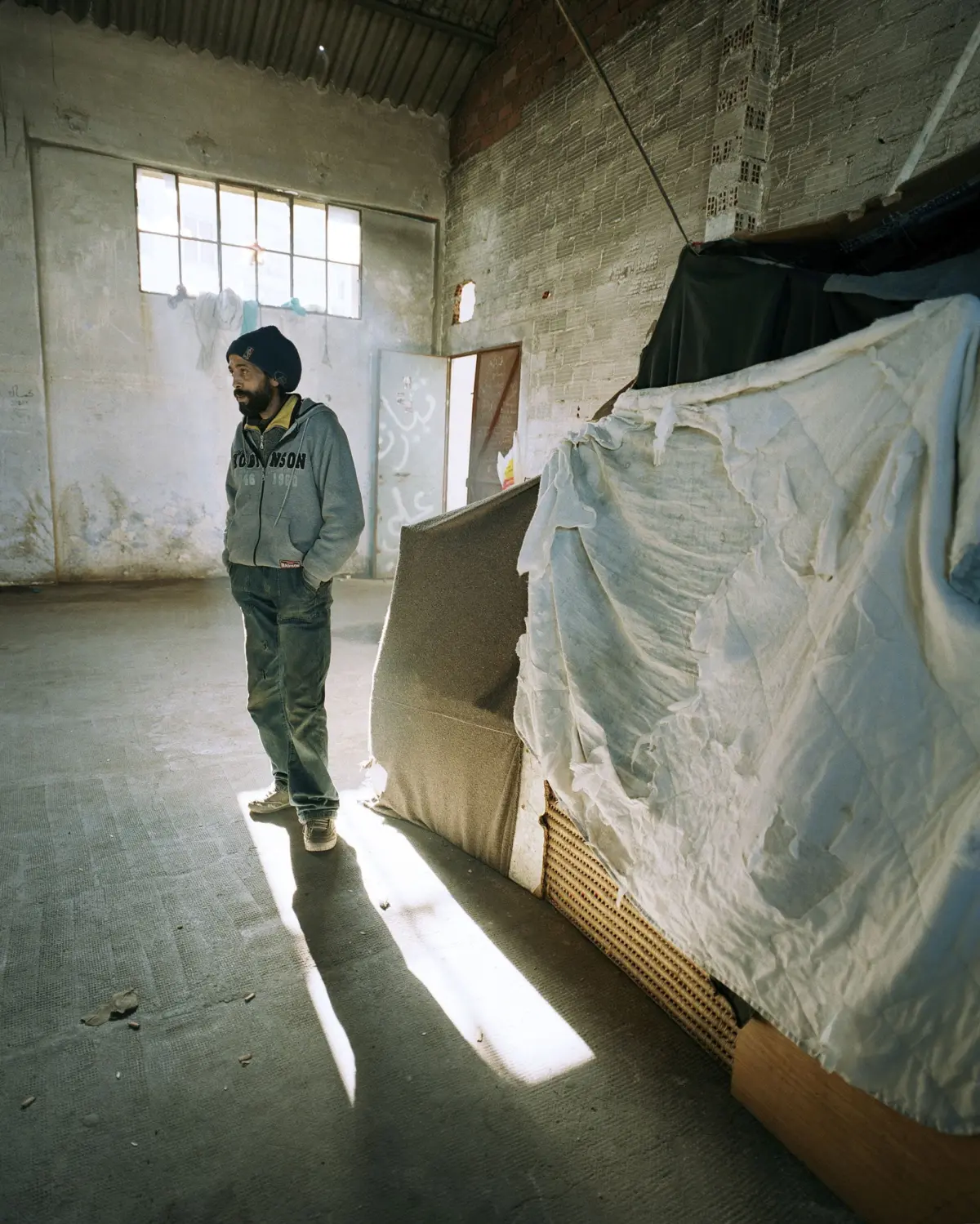 A person in a gray hoodie and beanie stands with hands in pockets in an abandoned warehouse, sunlit through a large window.