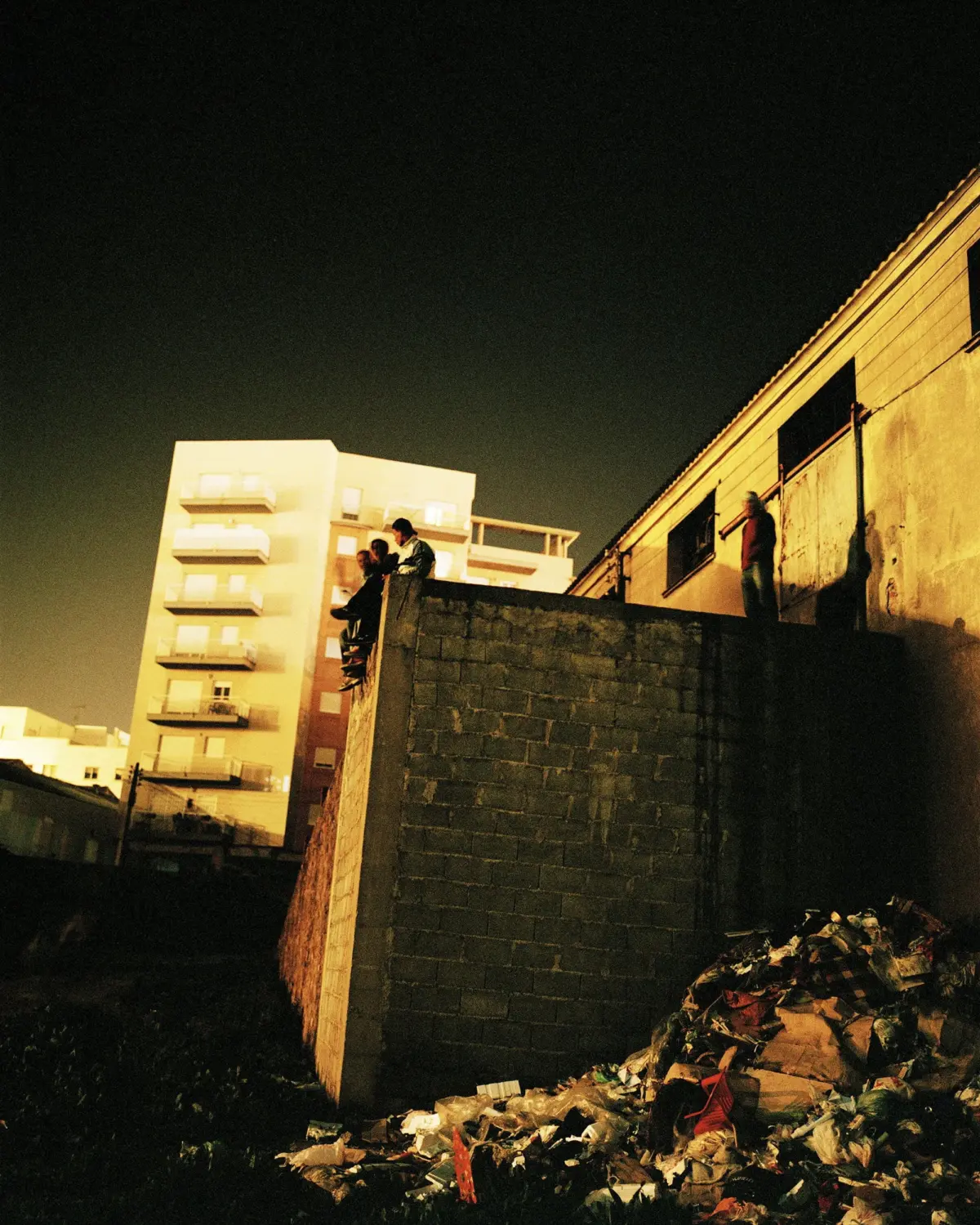 Group of people standing on a high concrete wall at night, overlooking a trash-strewn urban lot beside a building.